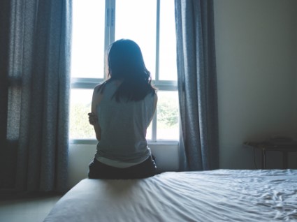 Woman sits on the corner of a bed looking out the window. 
