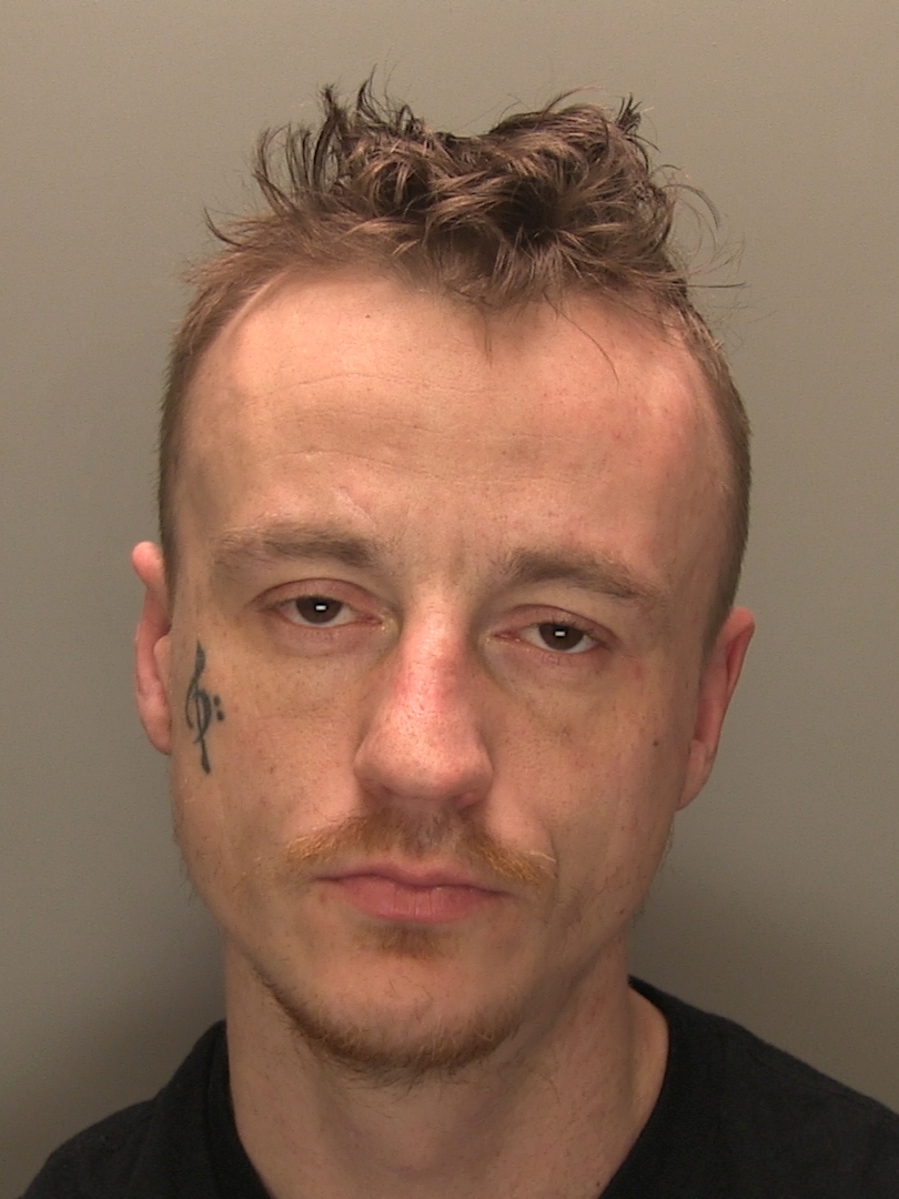 A mugshot of a man with short, spiky hair, a light moustache, and a bass clef tattoo on his right cheek.