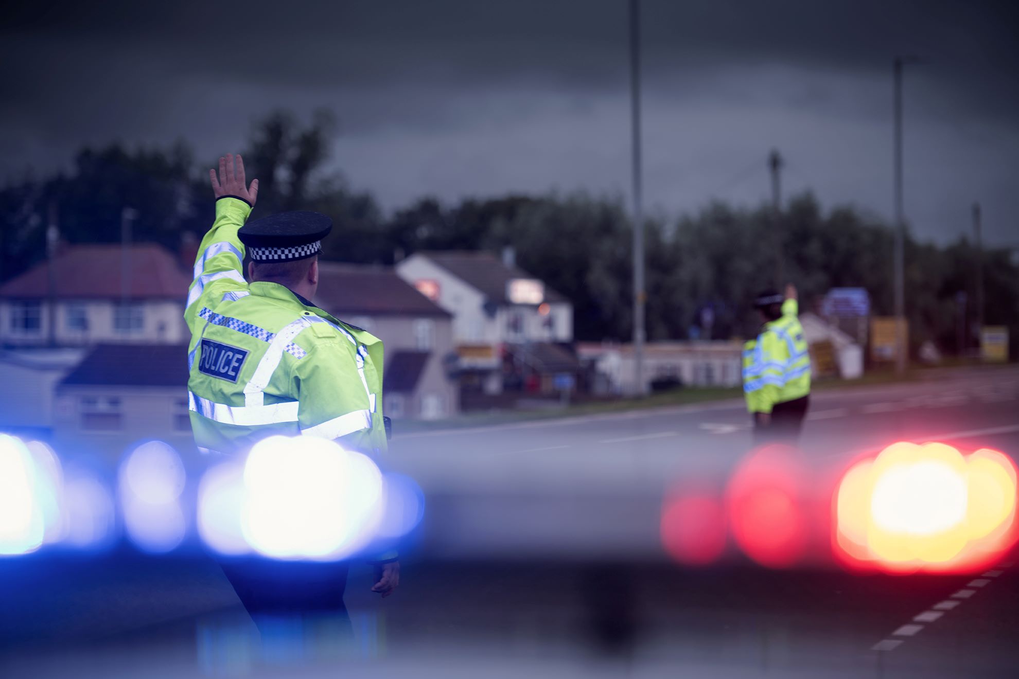 Two police officers with their back to the camera waving at cars during enforcement. Blue and red lights flash in the foreground.