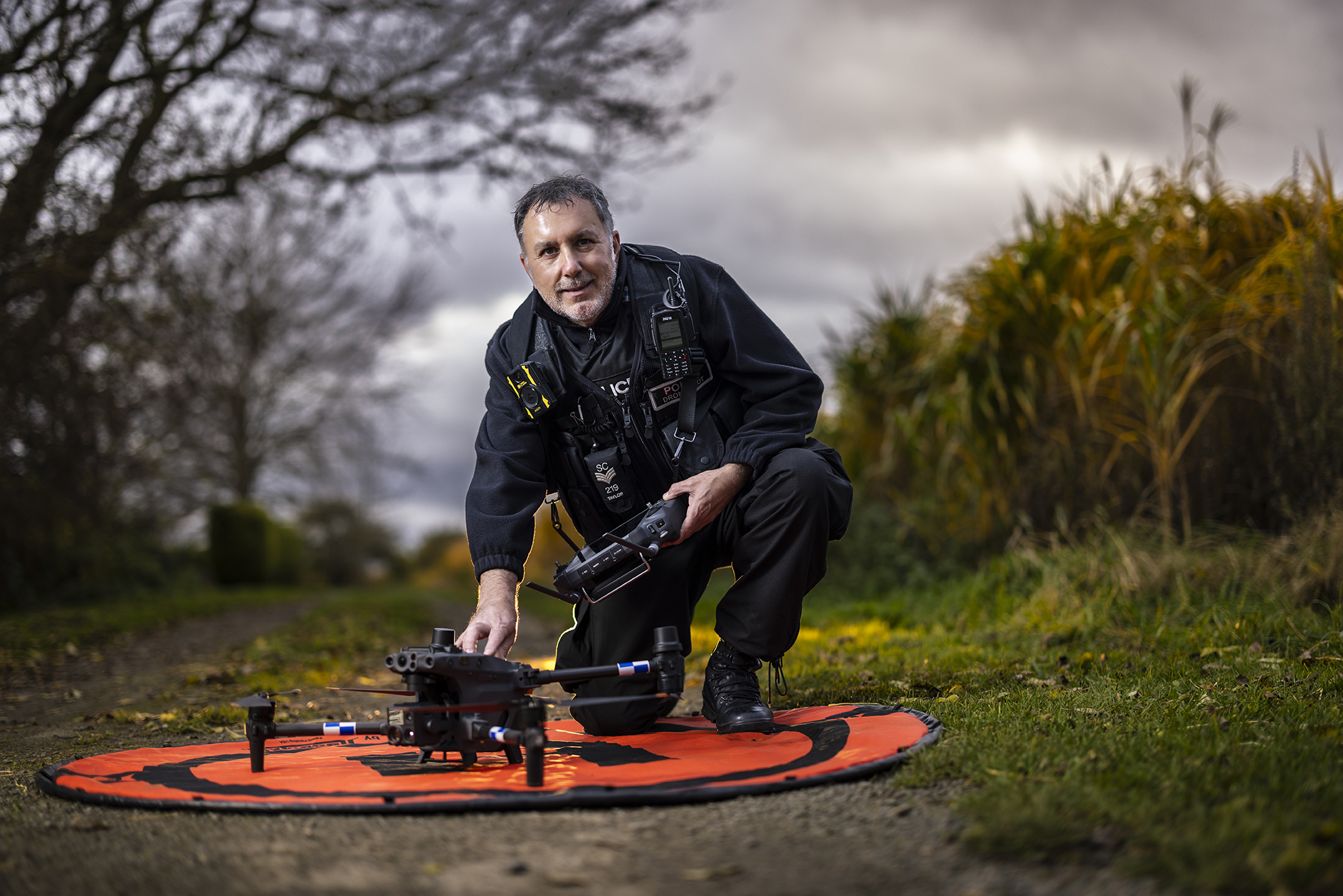 Chief drone pilot Kev Taylor. He is wearing dark uniform and is crouching on one knee, with a drone in front of him placed on an orange  and black mat. He is pictured in the countryside