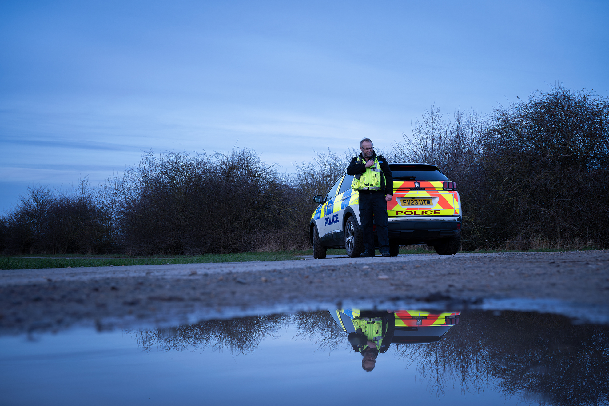 Muddy ground, puddle in the foreground, police officer and vehicle in the background