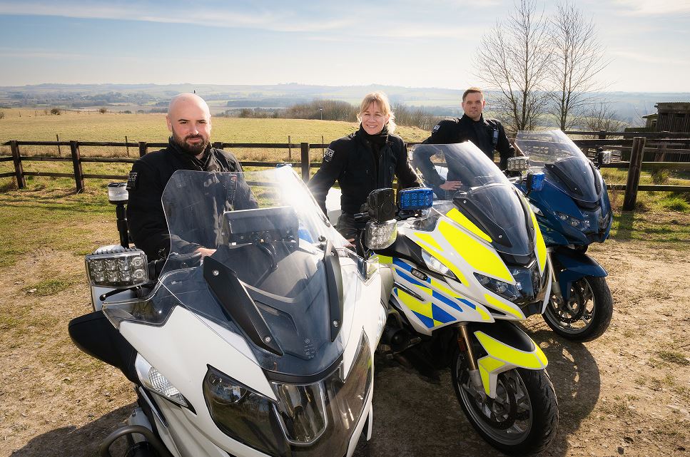 Three police motorcyclists sitting on two plain bikes and one marked bike (in the middle) with a field in the background. 