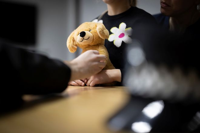 A small, fluffy teddy bear is held by a child, police hat is on the desk close up blurred in foreground