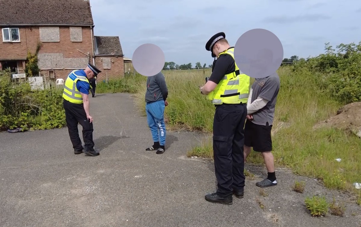 Two officers checking the papers of two men next to a house and field