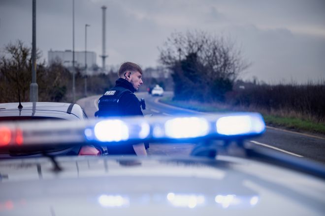 A police officer in a black uniform stands on the side of a road, viewed from behind a police car's emergency light bar.