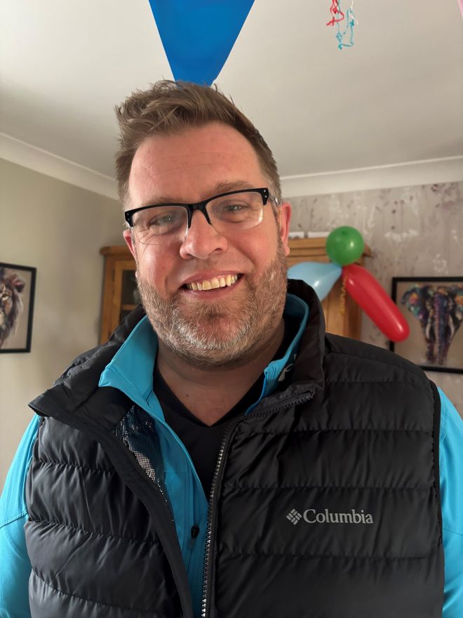 Man with a smiley face, short beard, wearing glasses and a black gillet with a light blue shirt