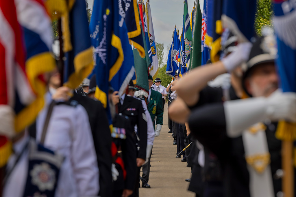 Rows of police officers holding ceremonial flags form a guard of honour along a pathway at an outdoor memorial event. The image is photographed with a shallow depth of field, with colourful blue, green and red standards framing the scene and a line of participants visible in the centre background.