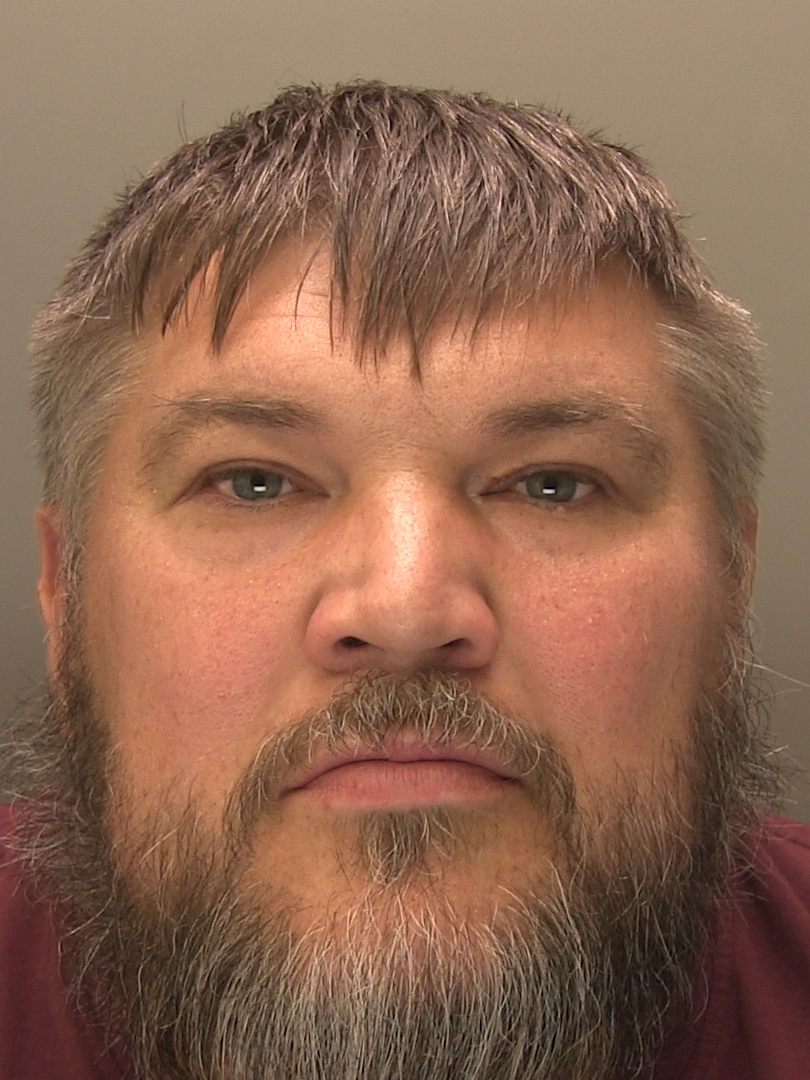 Head-and-shoulders portrait of a person facing the camera against a plain light-coloured background. The person has short hair with a fringe and a full beard with some grey colouring. The image is tightly cropped around the face and shoulders and is evenly lit.