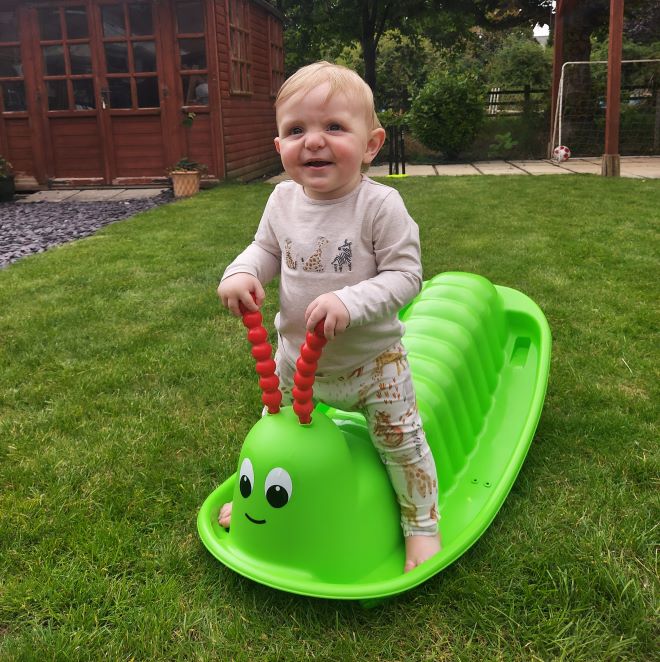 Photograph of a young boy with blonde hair sat on a green toy rocker in the garden