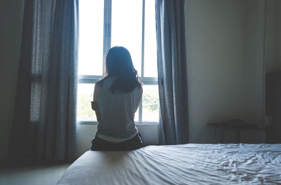 A woman with dark hair sits on the edge of a bed, facing a bright window with her arms crossed.