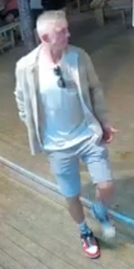 CCTV image of man with grey hair wearing a white top underneath a light coloured jacket. He has dark sunglasses hanging from his top and has red shoes.
