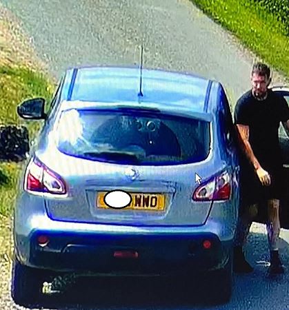A man in a black t-shirt stands by the open door of a silver Nissan Qashqai parked on a roadside.