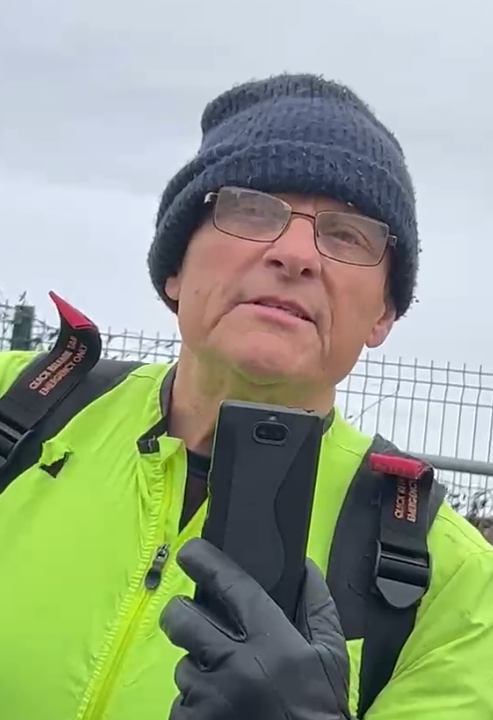 A man in a neon yellow high-visibility jacket and a dark beanie holds a smartphone while looking towards the camera.