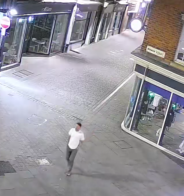 Man with dark coloured hair similar to a ‘mullet style’. He’s a white man, wearing a white t-shirt, grey colour jeans and trainers