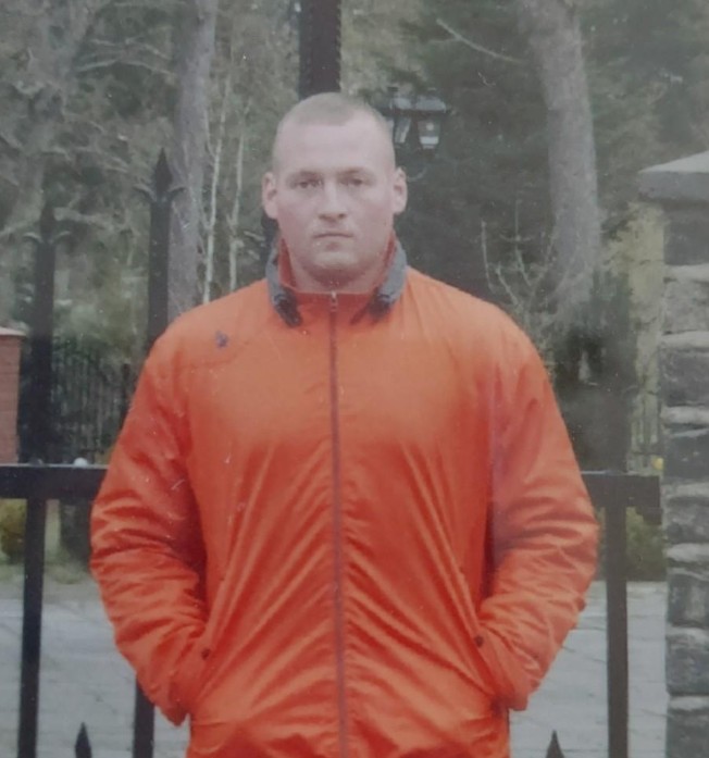 36-year-old Karol is looking at the camera and is wearing a bright orange jacket. He has his hands in his jacket pockets.