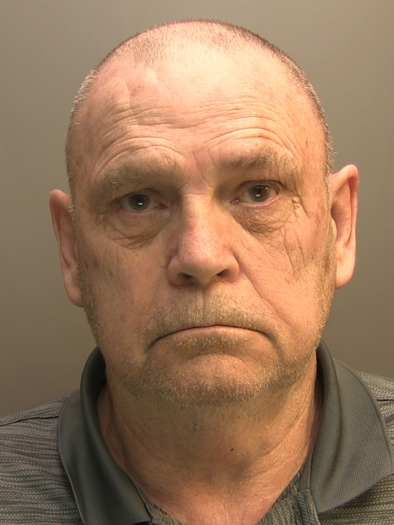 A mugshot of an older white man with a shaved head and grey stubble, wearing a grey polo shirt against a plain background.