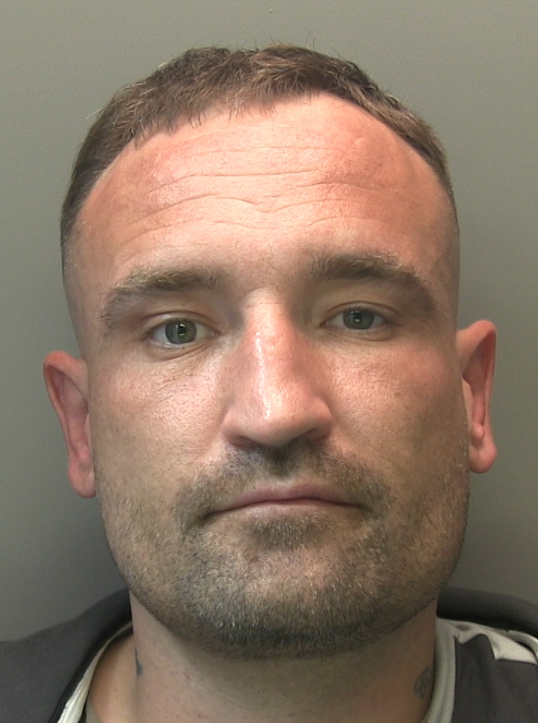 A head-and-shoulders mugshot of a man with short brown hair, light stubble, and a neutral expression.