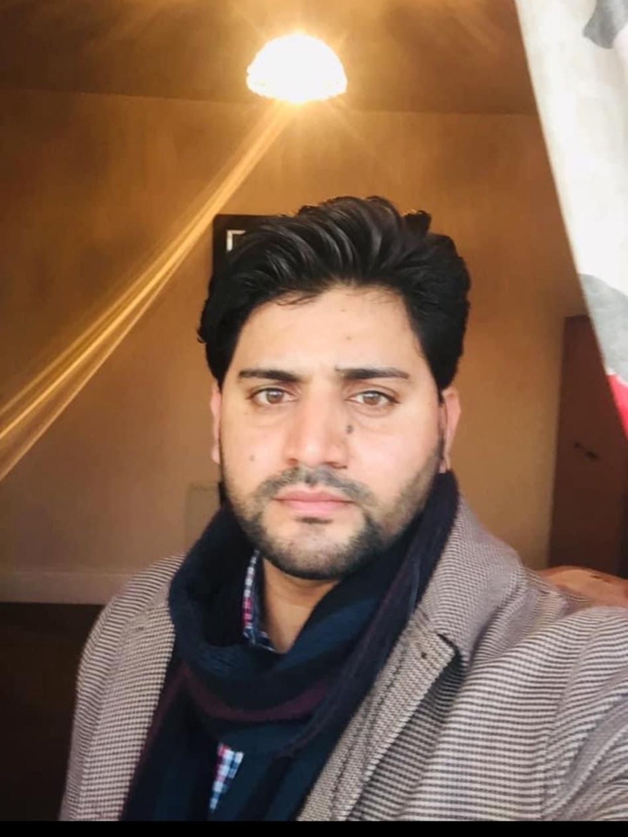 A photo of Shah Muhammad. He is wearing a coat and a scarf and is standing under a ceiling light. 